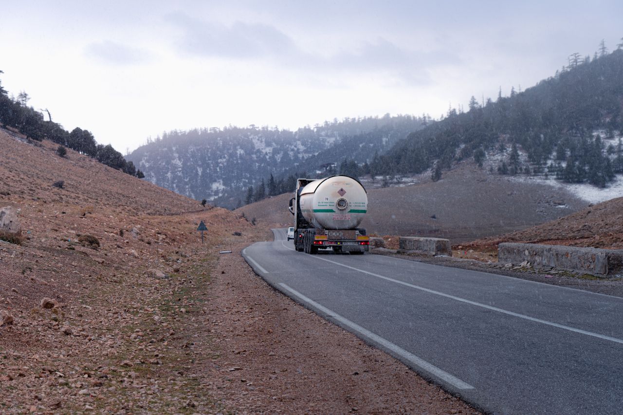 Gas tanker truck traveling on a mountainous road with snow-capped hills in the background.
