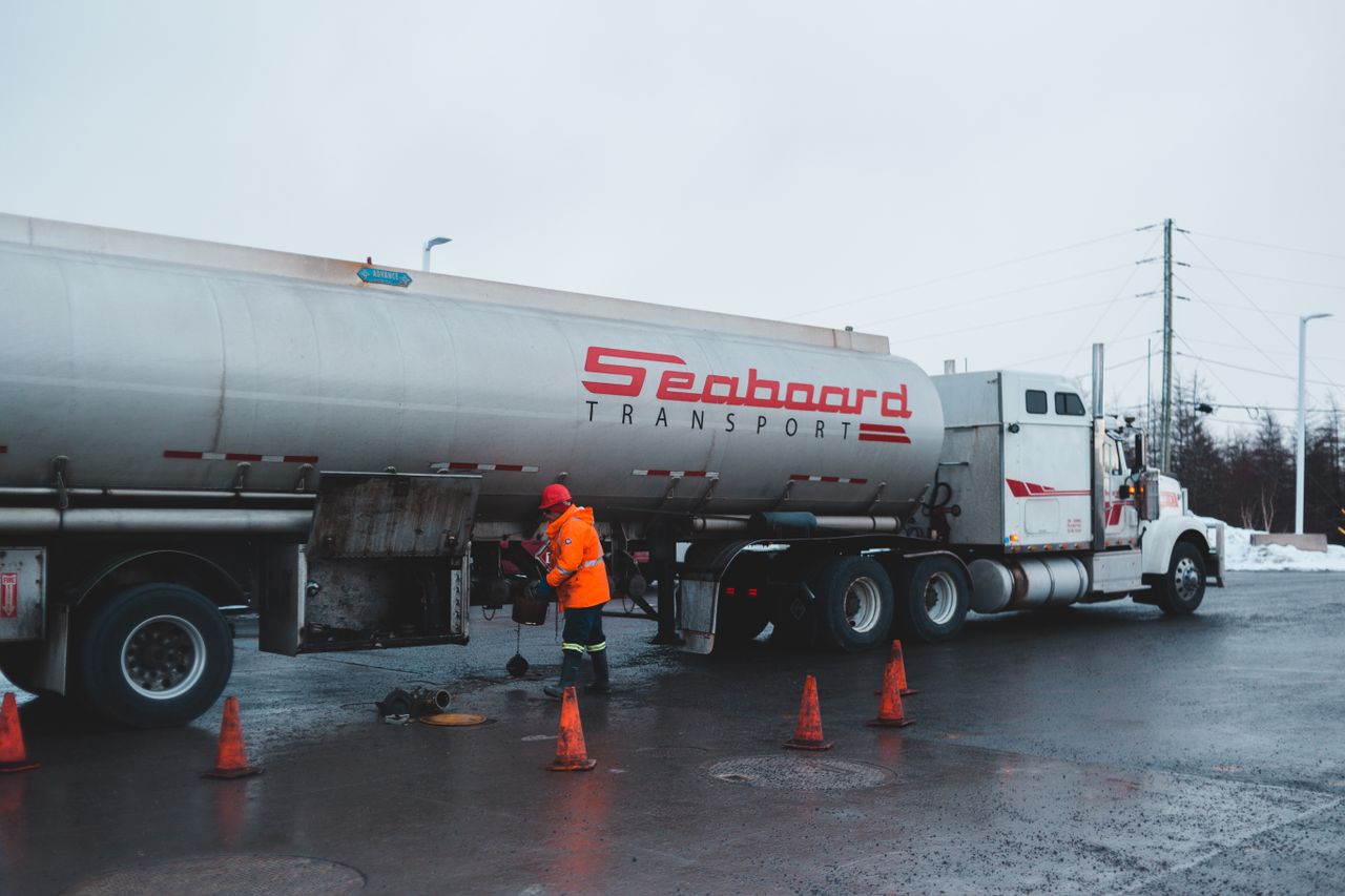 Back view of anonymous male worker in bright uniform and helmet cleaning oil truck vehicle while standing on asphalt roadway surrounded by barriers under white sky in town